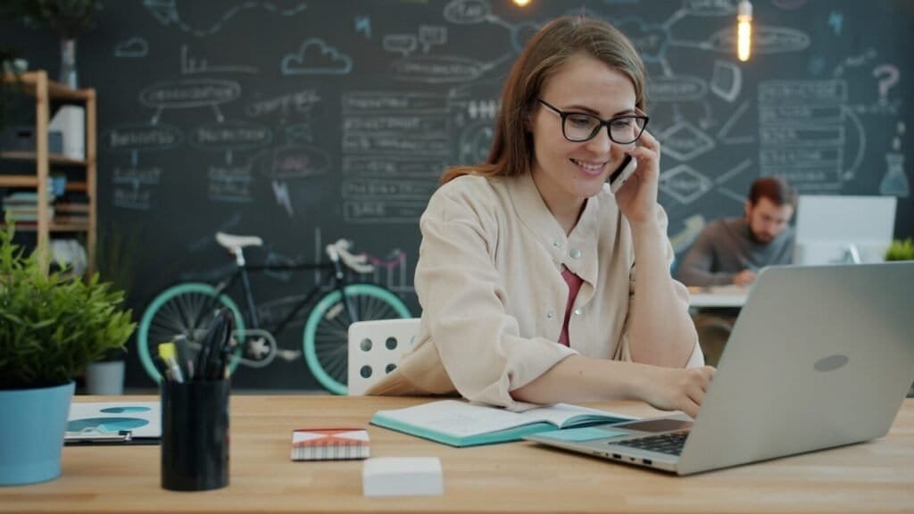 Woman Talking On Phone While Working On Laptop In Office